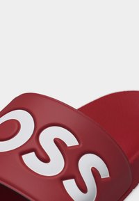 Κόκκινο σανδάλι με λαστιχένια σόλα και φαρδιά λουρί που φέρει μεγάλα λευκά ανάγλυφα γράμματα σε απλό ανοιχτό φόντο.