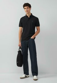 Schwarzes Poloshirt mit dezentem Logo, kombiniert mit dunklen Jeanshosen und weißen Sneakers mit schwarzem Riemen. Hält eine schwarze Tragetasche.