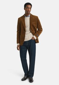 Man in een bruine corduroy blazer, crèmekleurige kabelgebreide trui, blauwe spijkerbroek en donkerbruine loafers, staand met een hand in zijn zak.