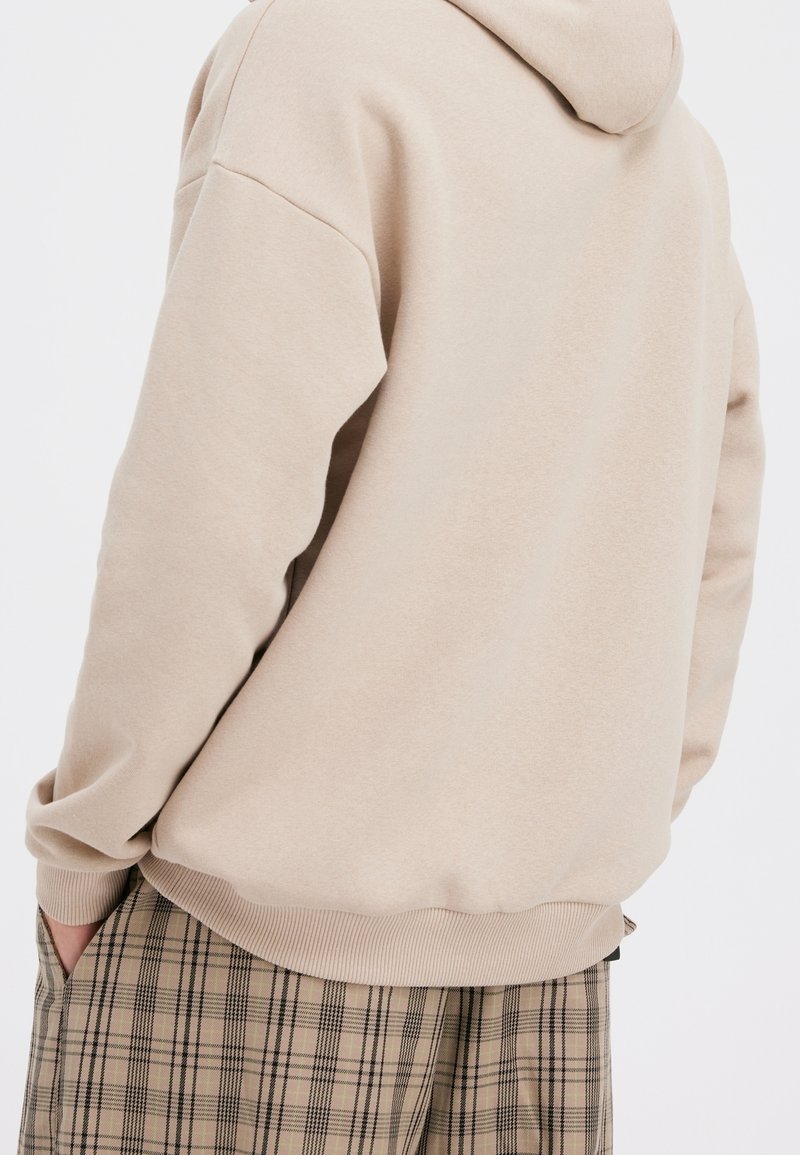 Beige huppari, jossa on pehmeä kangas, huppu ja resorit hihansuissa. Käytetty ruudullisten housujen päällä, rento malli ja arkinen tyyli.