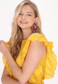 Abito giallo in tessuto a occhiello con maniche a volants, ritagli floreali e una finitura texturizzata. Grandi orecchini dichiarativi rossi e argento. Sfondo chiaro.
