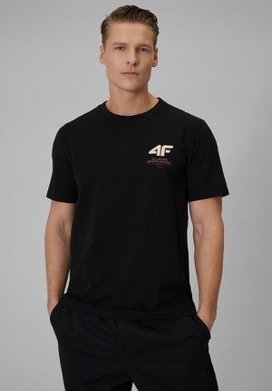 Młody mężczyzna z krótkimi włosami, ubrany w czarną koszulkę z logo "4G All-Round Sports Apparel", stoi z rękami w kieszeniach, neutralne tło.