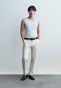 Ung mand står med hænderne i lommerne iført lysegrå tanktop, beige bukser, sort bælte og sorte støvler mod en ensfarvet baggrund.