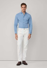 Hombre de pie con una camisa de botón azul claro, jeans blancos y botas marrón oscuro, frente a un fondo claro y liso.