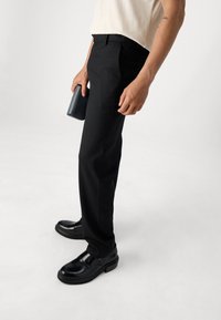 Schwarze maßgeschneiderte Hosen, glatter Stoff, gerader Schnitt, ergänzt mit einer dunklen Geldbörse. Getragen mit polierten schwarzen Schuhen.