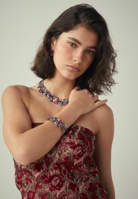 Bunte Edelstein-Schmuckstücke, bestehend aus einer Halskette und einem Armband mit verschiedenen Formen. Das Model trägt ein trägerloses Oberteil mit floralem Muster.