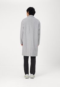 Cappotto di lana grigio con taglio dritto, maniche lunghe e colletto a punte, abbinato a pantaloni scuri e sneakers bianche.