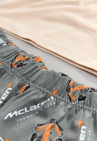 Grå stofshorts med et helt dækkende mønster af McLaren Formel 1 i orange og sort, med en elastisk talje. En lys beige top er bagved.