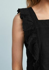 Sort linnedragt med firkantet udskæring og en flæset kant på skulderen. Stoffet har en tekstureret overflade.