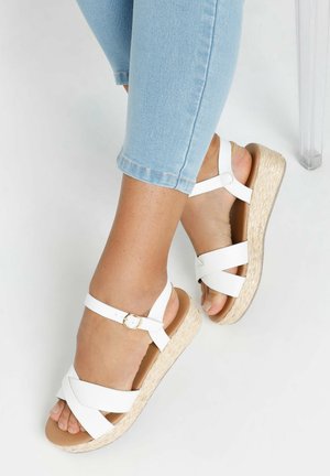 Sandalias de cuña blancas con correas entrecruzadas, hebilla ajustable en el tobillo y suela de cuña con textura de yute natural, combinadas con jeans de un azul claro.