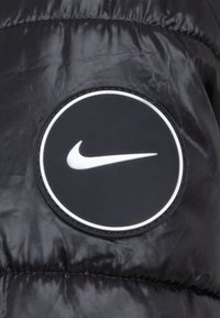 Μαύρο μπουφάν από πούπουλο με υλικό που έχει μια στρογγυλή, μαύρη ετικέτα με το λευκό λογότυπο Nike. Λείο υλικό με ραμμένες περιοχές.