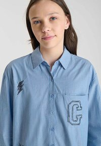 Light blue striped button-up shirt with a collar, featuring a stitched lightning bolt and a pocket with an embroidered "G."