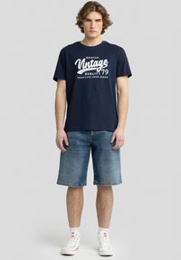 Tamsiai mėlyna medvilninė marškinėliai su baltu vintage stiliaus grafiniu spaudiniu, derinami su džinsiniais šortais ir baltais aukštakulnių sportbačiais. Kasdieniška, laisva fasonas.