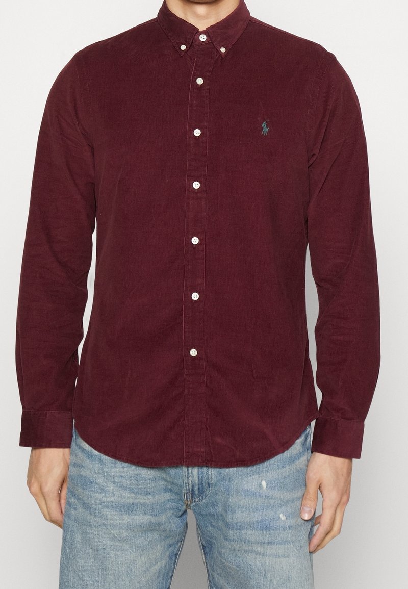 Dlouhý rukáv z manšestru v barvě burgundské s límečkem na knoflíky, zdobený malým zeleným logem na prsou a bílými knoflíky.