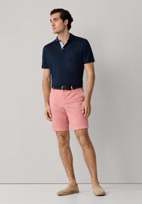 Homme portant un polo bleu marine, un short rose avec une ceinture foncée, et des chaussures beiges sans lacets, debout devant un fond uni gris clair.