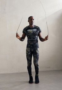 Man in black patterned athletic wear skipping rope against plain light wall in indoor setting.