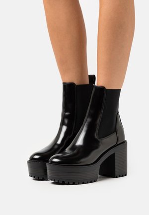 Černé kotníkové boty z lakované kůže s elastickými bočními panely, robustními blokovými podpatky a texturovanou gumovou podrážkou pro lepší trakci a stabilitu.