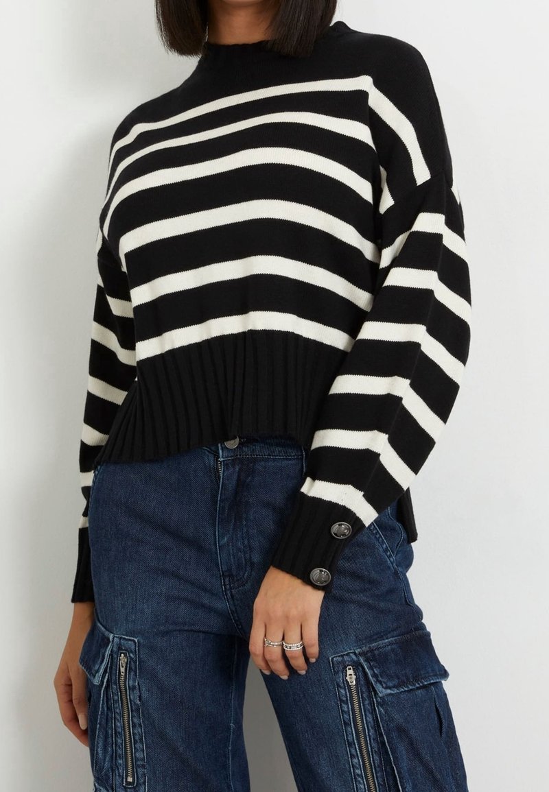 Maglione a righe bianche e nere realizzato a maglia, con orlo a coste, scollatura rotonda e dettagli a bottone sulle maniche, abbinato a pantaloni cargo in denim scuro.