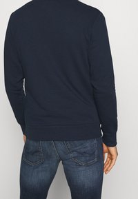 Man med mörkblå sweatshirt och blekblå jeans, sedd bakifrån mot en enfärgad ljus bakgrund.