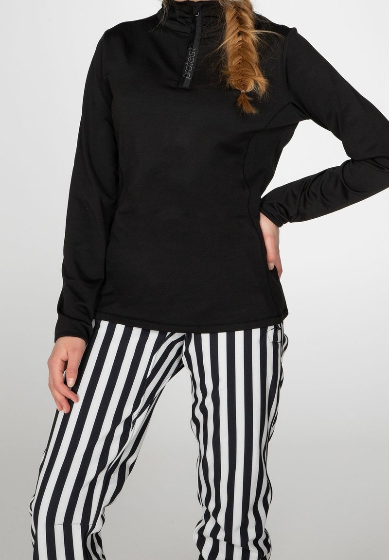 Kvinna iklädd en svart långärmad topp och byxor med svart-vita vertikala ränder, med en fiskbensfläta över axeln.