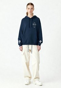 Felpa con cappuccio blu navy con tasca frontale e grafica a croce bianca. Abbinata a pantaloni cargo crema e sneakers bianche.