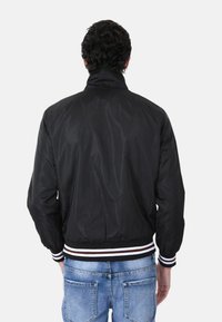 Giacca bomber nera realizzata in tessuto liscio con colletto alto. Caratterizzata da un orlo e polsini a coste a righe, abbinata a jeans blu chiaro.