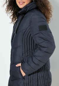 Žena s kudrnatými vlasy, oblečená v tmavé zateplené bundě se zapínáním na zip a bočním prošíváním, má ruce v kapsách a límec se stahovacími šňůrkami.