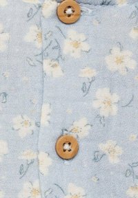 Svijetloplavi materijal s cvjetnim uzorkom, koji sadrži bijele i žute cvjetove. Drveni gumb s izrezbarenim dizajnom je istaknut.