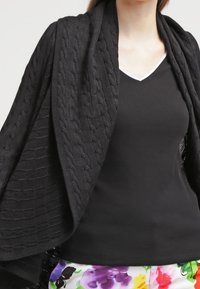 Woman wearing a black cable-knit shawl over a black V-neck top and colorful floral pants.