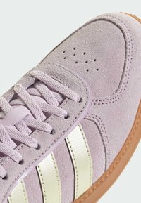 Fioletowy zamszowy sneaker z trzema metalicznymi srebrnymi paskami, teksturowanymi sznurowadłami i gumową podeszwą. Na obszarze noska znajdują się perforacje.