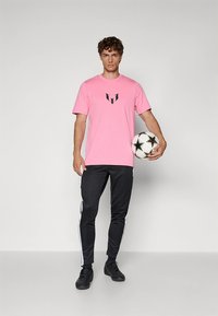 Rosa kurzärmeliges T-Shirt mit schwarzem Logo auf der Brust, kombiniert mit schwarzen Jogginghosen mit weißen Seitenstreifen. Hält einen Fußball.