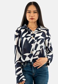 Camisa corta de botones en azul marino y blanco con patrones abstractos, de mangas largas y con collar. Combinada con jeans azules.