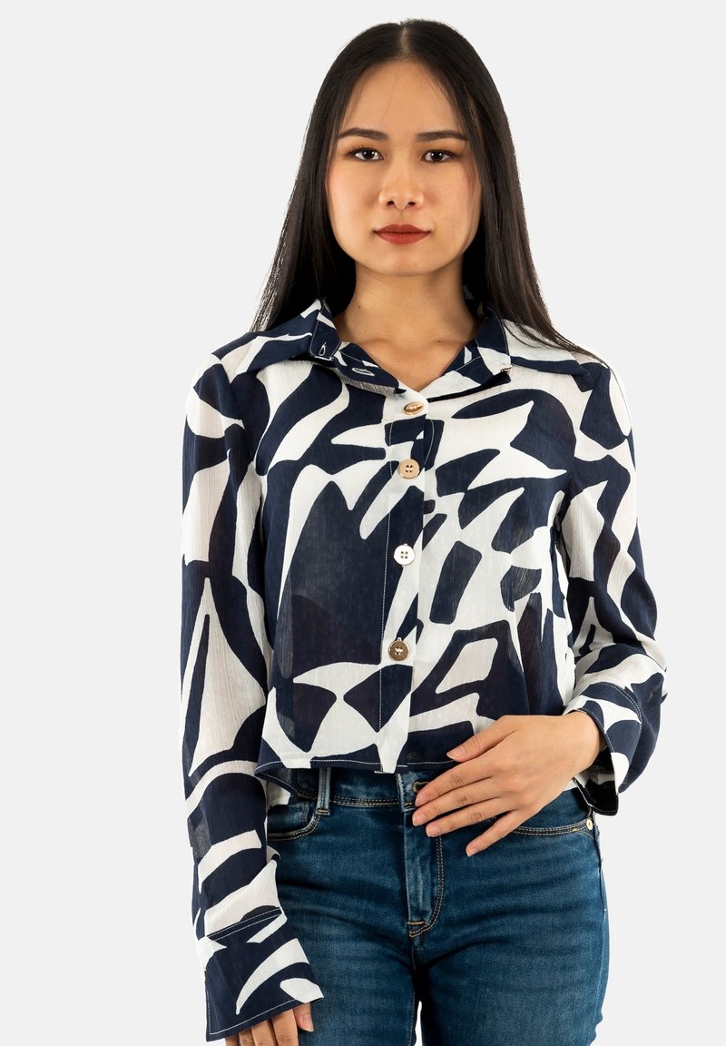 Camisa corta de botones en azul marino y blanco con patrones abstractos, de mangas largas y con collar. Combinada con jeans azules.