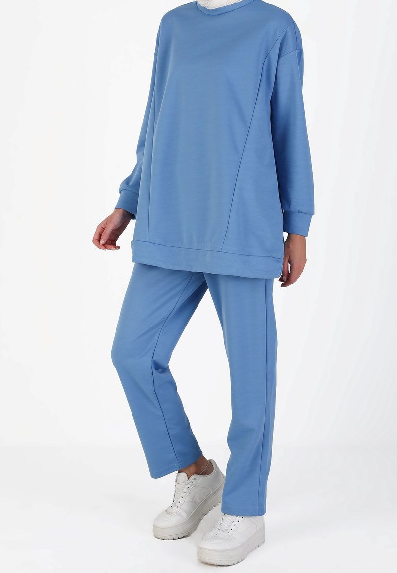 Personne portant un haut bleu ample à manches longues et un pantalon assorti, associés à des baskets blanches à plateforme, debout devant un fond uni.