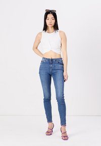 Vitt knutet crop-top, blå skinny jeans och lila sandaletter med remmar. Modellen poserar mot en enfärgad vit bakgrund.