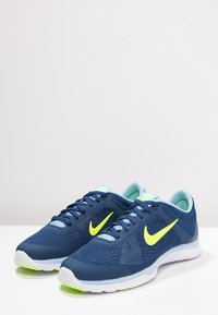 Scarpe da ginnastica blu navy con tomaia in rete, swoosh giallo neon, dettagli azzurri e suola bianca. Presentano un design con lacci e sovrapposizioni testurizzate.