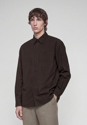 Junger Mann steht vor einem einfarbigen Hintergrund, trägt ein dunkelbraunes Hemd mit Knöpfen und karierte Hose, eine Hand in der Tasche.