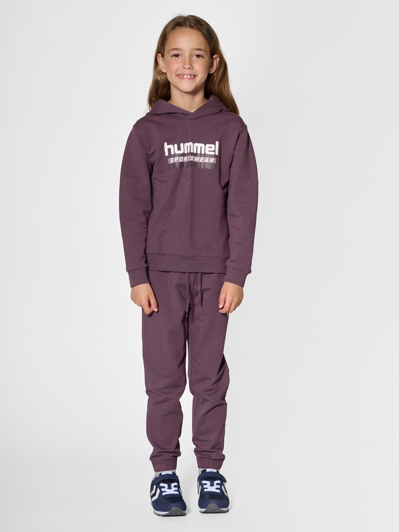 Jong meisje staat, draagt een bijpassende paarse hummel sporthoodie en joggingbroek met blauwe sneakers, glimlachend naar de camera.