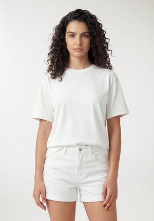 Giovane donna con capelli ricci e scuri, indossa una maglietta bianca a maniche corte semplice e pantaloncini bianchi con orlo arrotolato, in piedi su sfondo bianco.