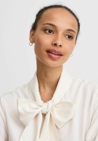 Blouse en crème avec un grand nœud au cou. Texture lisse avec un design drapé, mettant en valeur un style décontracté et élégant.