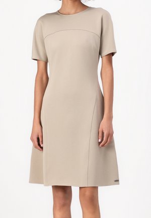 Mujer vistiendo un vestido beige hasta la rodilla, de manga corta, con escote redondo y detalles sutiles en las costuras.