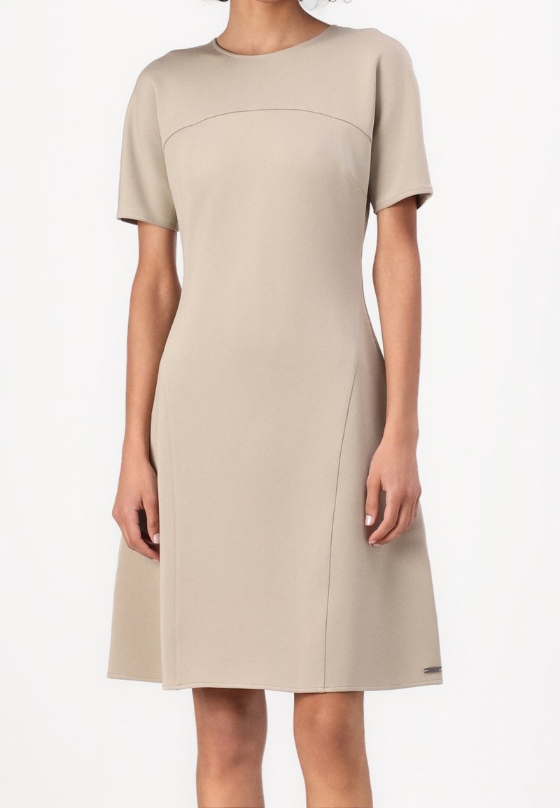 Femme portant une robe beige, longueur genou, manches courtes, avec un col rond et des détails discrets de couture.
