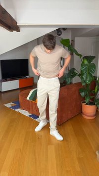 Man adjusting light beige pants in living room with brown couch, large green plant, flat-screen TV, and wooden floor.