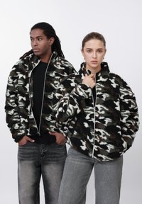 Fleece jakker med lynlås i et camouflagemønster i sort, hvid og olivengrøn. Begge har høje kraver og er stylet med grå jeans.