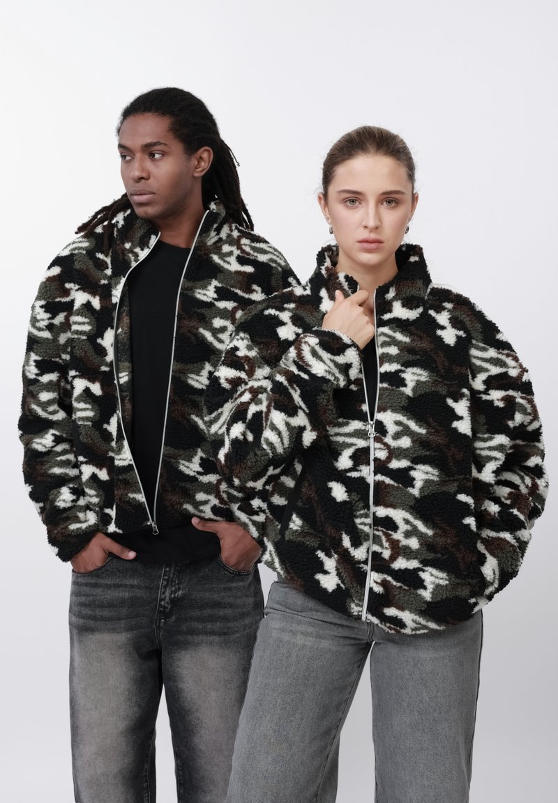 Fekete, fehér és olíva terepmintás, polár zipzáras dzsekik. Mindkettő magas nyakkal rendelkezik és szürke farmerrel van párosítva.