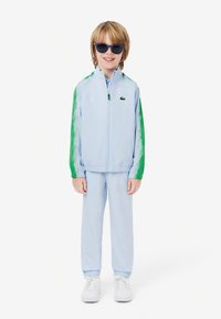 Jongentje in een lichtblauw trainingspak met groene strepen op de mouwen, witte sneakers en donkere zonnebril, staand en lachend.