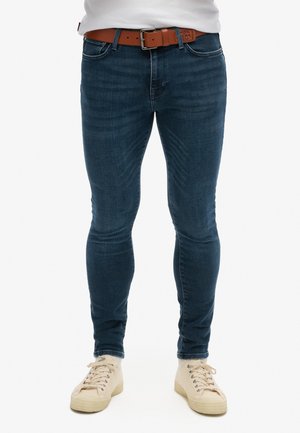 Jeans de denim azul de corte skinny con un ligero desvanecimiento, diseño de cinco bolsillos y un cinturón de cuero marrón. Combinados con zapatillas de lona beige claras.
