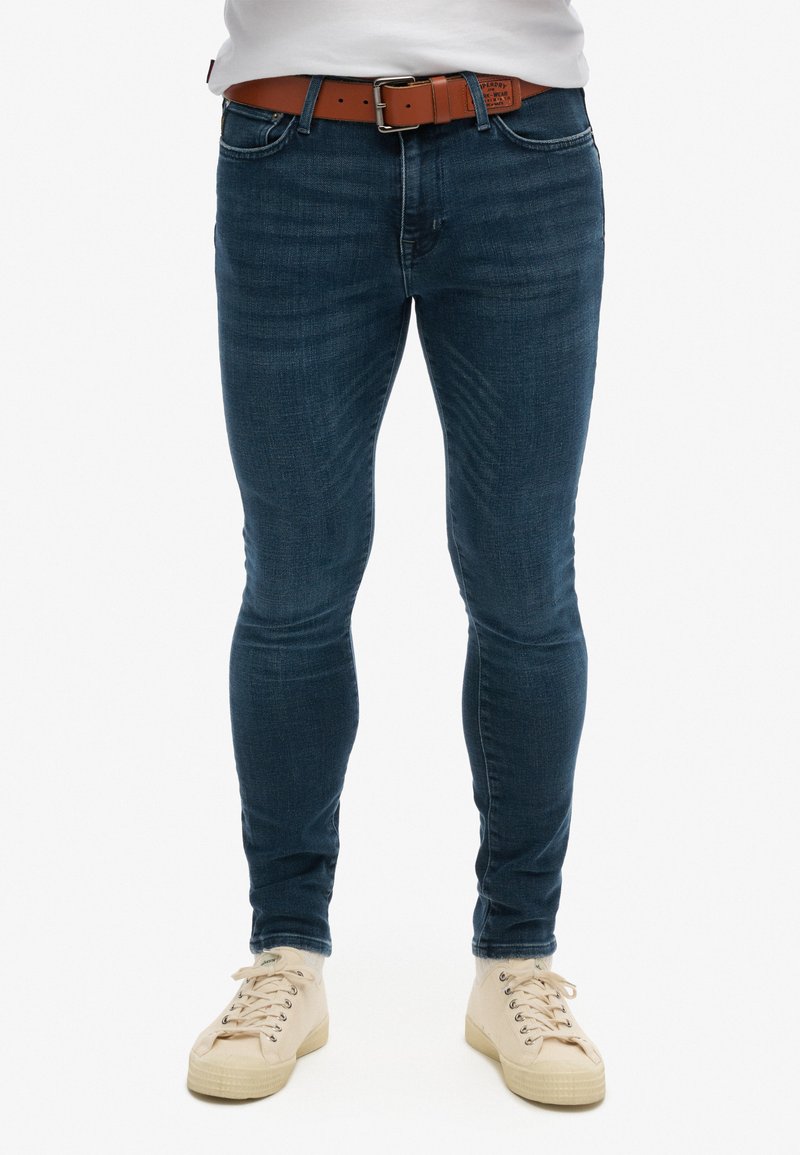 Jeans de denim azul de corte skinny con un ligero desvanecimiento, diseño de cinco bolsillos y un cinturón de cuero marrón. Combinados con zapatillas de lona beige claras.