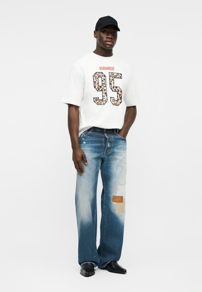 Μαύρος άνδρας που φοράει λευκό T-shirt DSQUARED2 με τον αριθμό "95", τζιν με μπλε μπαλώματα, μαύρα παπούτσια και μαύρο καπέλο, όρθιος με το ένα χέρι στην τσέπη.