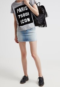 Person som bär en grå "PARIS 75001 ICON" t-shirt, en denim minikjol, svarta skor och bär en svart fransryggsäck.